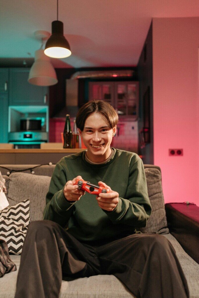 A young man happily playing video games on a couch in a cozy and colorful living room.