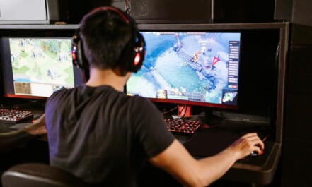 Adult male gamer immersed in PC gaming on dual monitors with headphones indoors.