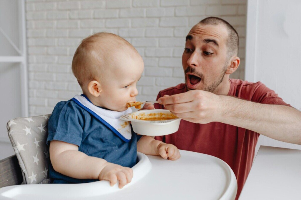 Loving father feeds baby in a high chair, bonding moment indoors.