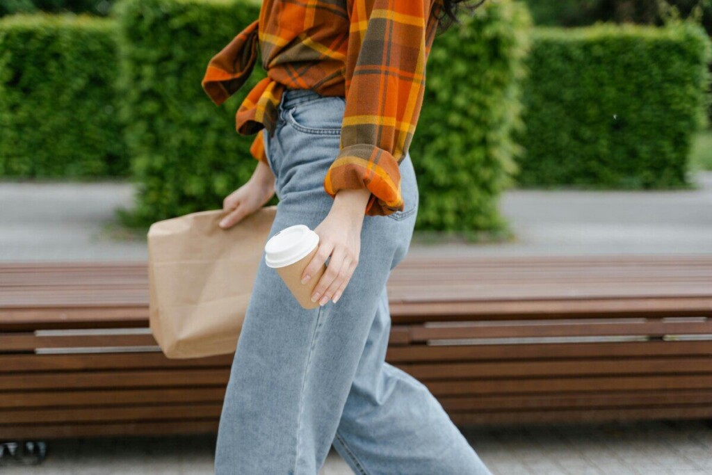 Woman walking outdoors holding a coffee cup and brown paper bag, relaxed lifestyle.