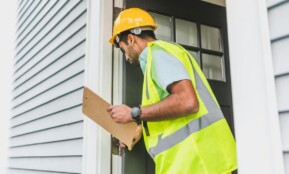 A male inspector in a hard hat conducts a property inspection outdoors with a clipboard.