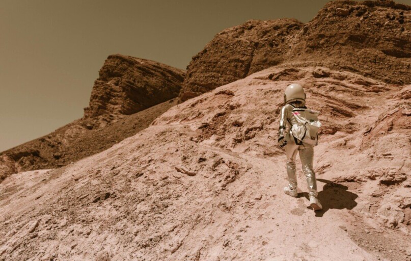 Astronaut in a space suit walking uphill on a rocky, desert-like alien planet.
