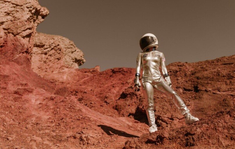 Astronaut in space suit standing on red rocky terrain resembling Mars' surface.
