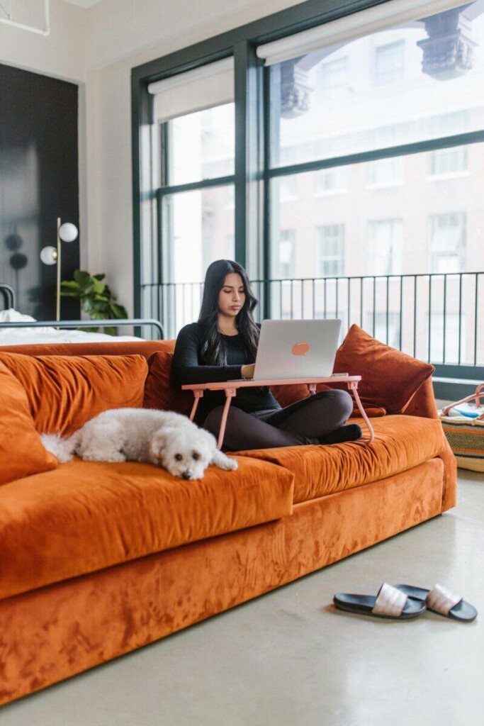 A woman works on her laptop at home with a dog beside her on an orange sofa.