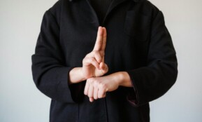 A person in a black coat demonstrating a sign language gesture indoors.