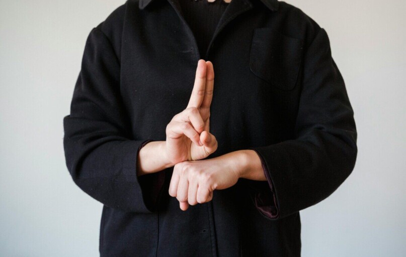 A person in a black coat demonstrating a sign language gesture indoors.