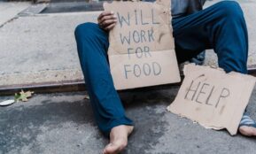 A homeless person sitting on the street holding a cardboard sign with the message 'Will work for food'.