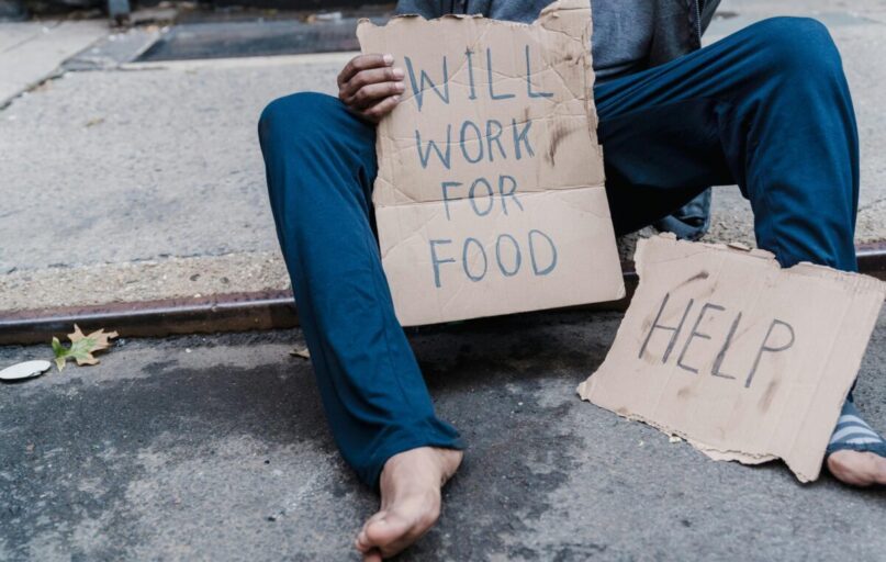 A homeless person sitting on the street holding a cardboard sign with the message 'Will work for food'.