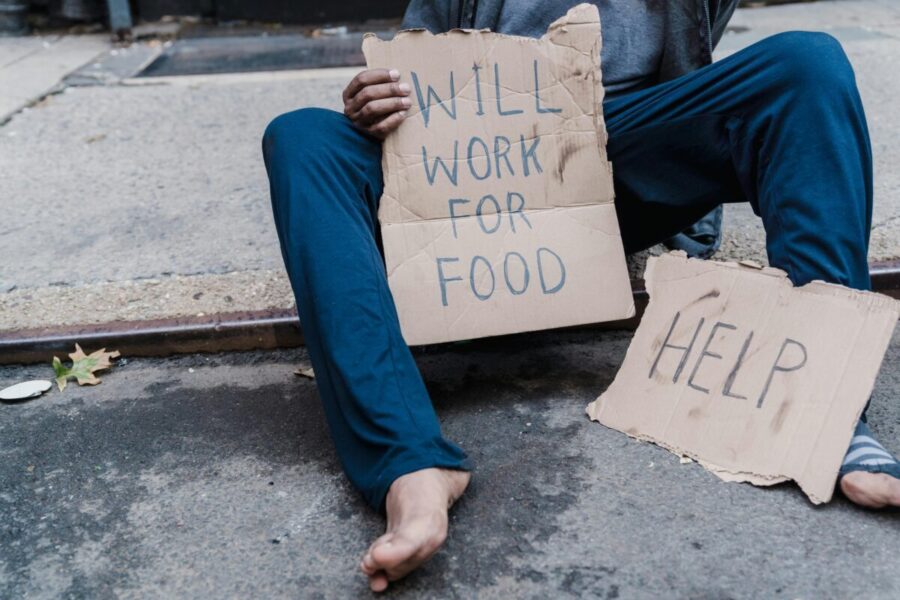 A homeless person sitting on the street holding a cardboard sign with the message 'Will work for food'.
