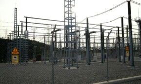 Outdoor electrical power substation with high voltage equipment and safety signs, surrounded by fencing.