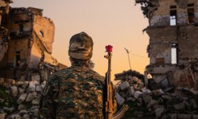 A soldier in camouflage with a red rose on rifle amidst ruined buildings at sunset.