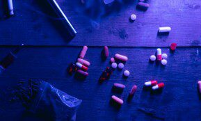 A flat lay of various drugs including pills, capsules, and a syringe on a wooden surface under moody blue lighting.