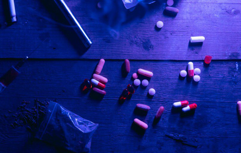 A flat lay of various drugs including pills, capsules, and a syringe on a wooden surface under moody blue lighting.