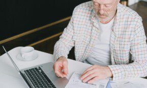 Elderly man with eyeglasses reviewing documents at a laptop. Indoor setting with natural light.