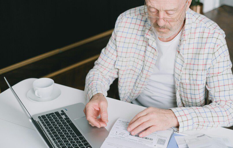 Elderly man with eyeglasses reviewing documents at a laptop. Indoor setting with natural light.