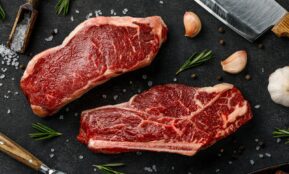 two steaks on a cutting board next to garlic, garlic, and a knife