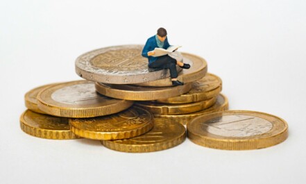 Miniature person sitting on stack of coins reading newspaper