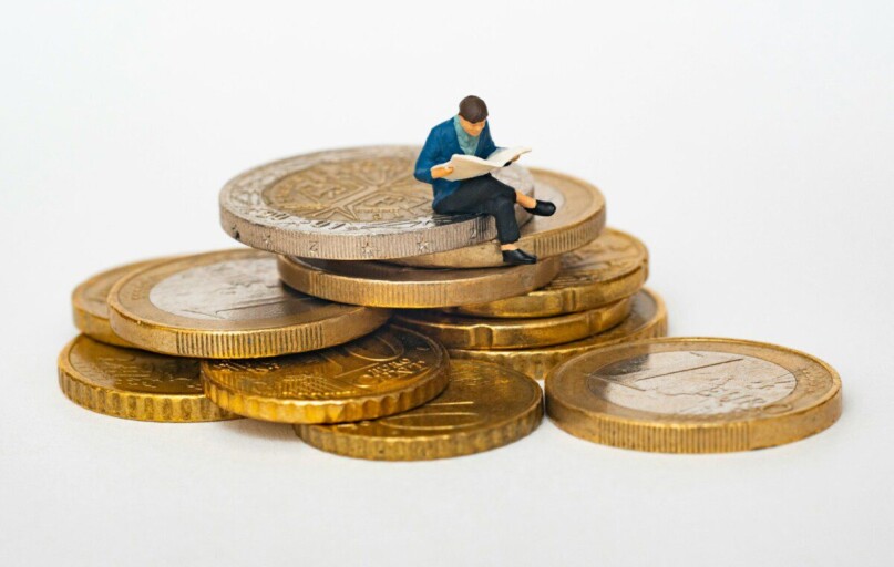 Miniature person sitting on stack of coins reading newspaper