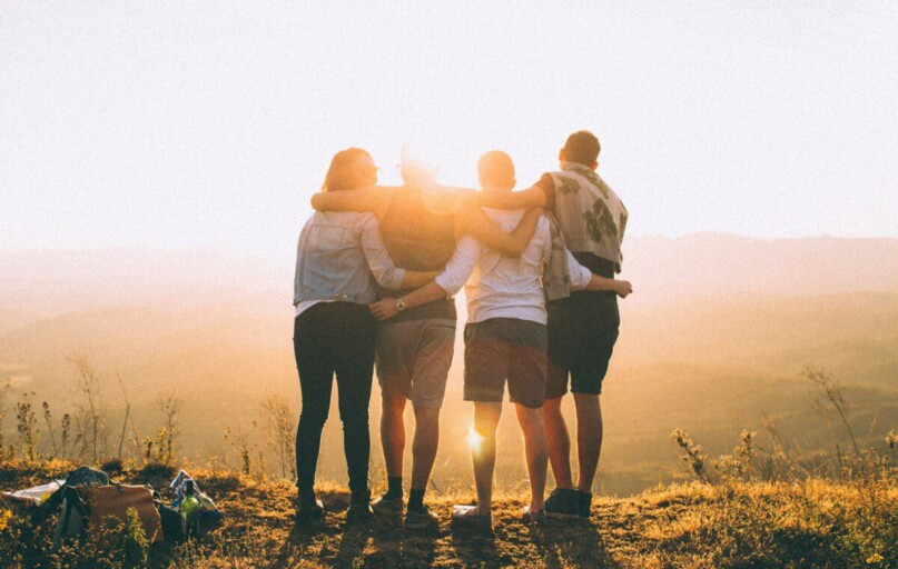 four person hands wrap around shoulders while looking at sunset