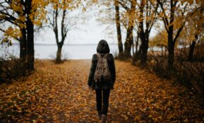 woman walking on pathway with falling leaves near body of water during daytime