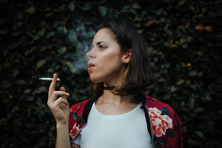 Portrait of a woman smoking outdoors, clad in a floral jacket, exhaling smoke against a leafy backdrop.