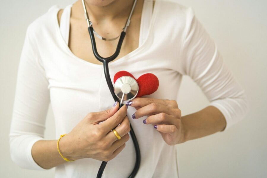 Unrecognizable female wearing white shirt while standing on white background with diaphragm of stethoscope on red handmade heart in room
