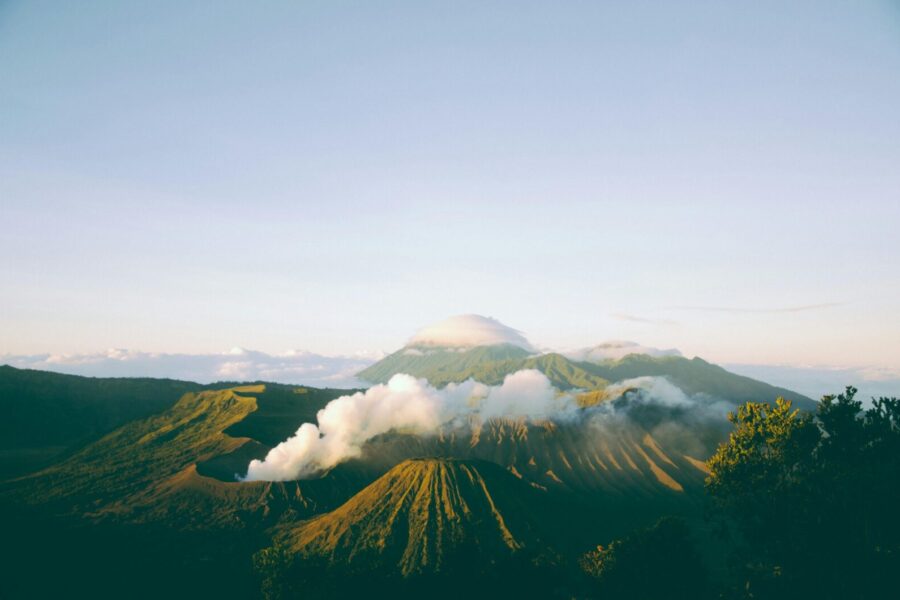 A breathtaking view of Mount Bromo emitting smoke at sunrise, surrounded by lush landscapes of East Java, Indonesia.