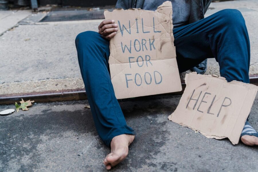 A homeless person sitting on the street holding a cardboard sign with the message 'Will work for food'.