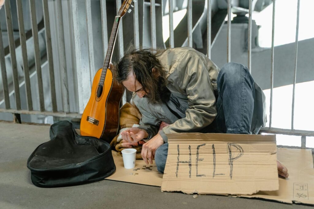A homeless man sits by a railing with a cardboard sign reading 'HELP', coins, and a guitar.